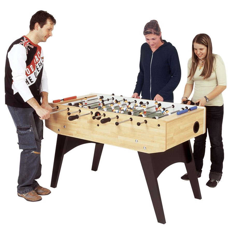 Drei Personen spielen Tischfußball an einem Holztisch mit schwarzen Beinen, konzentriert auf das Spiel.