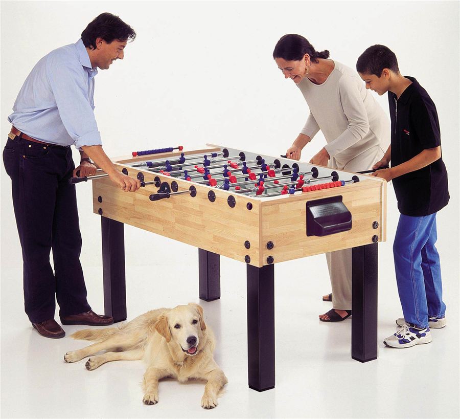 Drei Personen spielen Tischfußball, während ein Hund entspannt auf dem Boden liegt.