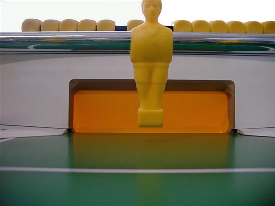 Nahaufnahme einer gelben Kickerfigur vor einem Tor auf einem grünen Spielfeld eines Tischfußballspiels.