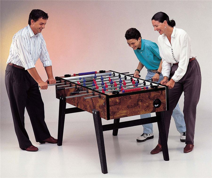Drei Personen spielen lachend Tischfußball an einem Holztisch mit bunten Spielfiguren.