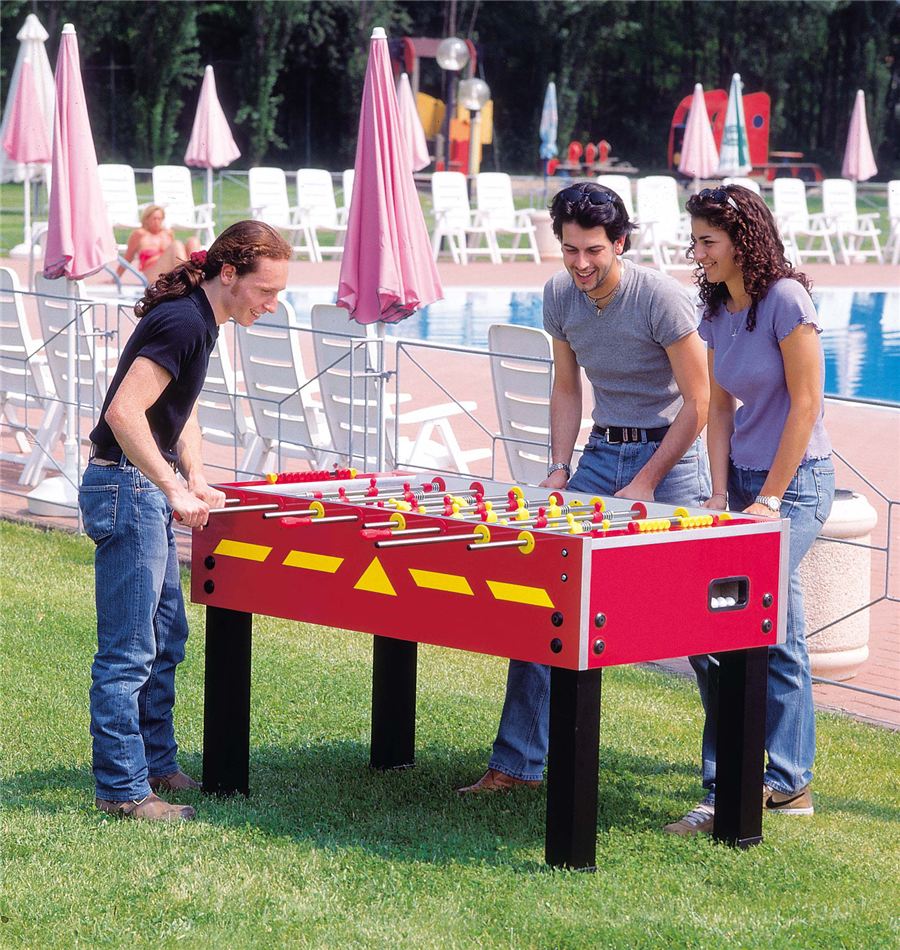 Drei junge Erwachsene spielen lachend an einem roten Kickertisch im Freien neben einem Pool mit Liegestühlen und Sonnenschirmen.