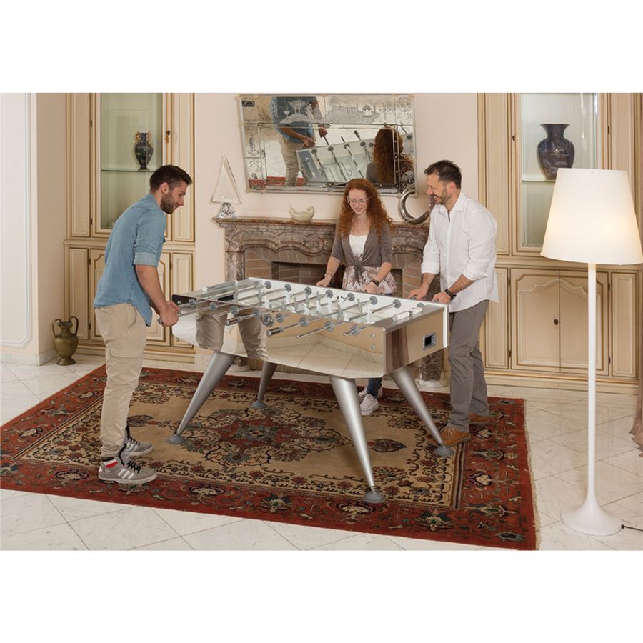 Drei Personen spielen Tischfußball in einem elegant eingerichteten Wohnzimmer mit Kamin und Teppich.