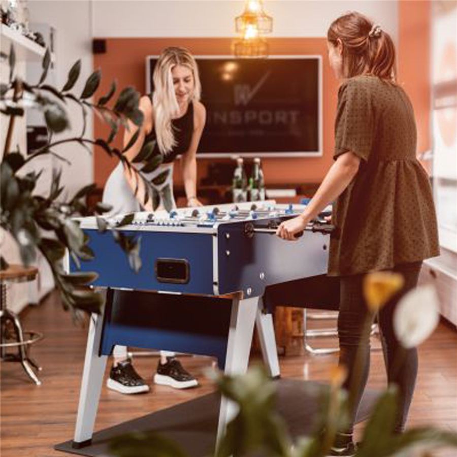 Zwei Frauen spielen an einem blauen Kickertisch in einem modern eingerichteten Raum mit Pflanzen und Flachbildfernseher im Hintergrund.